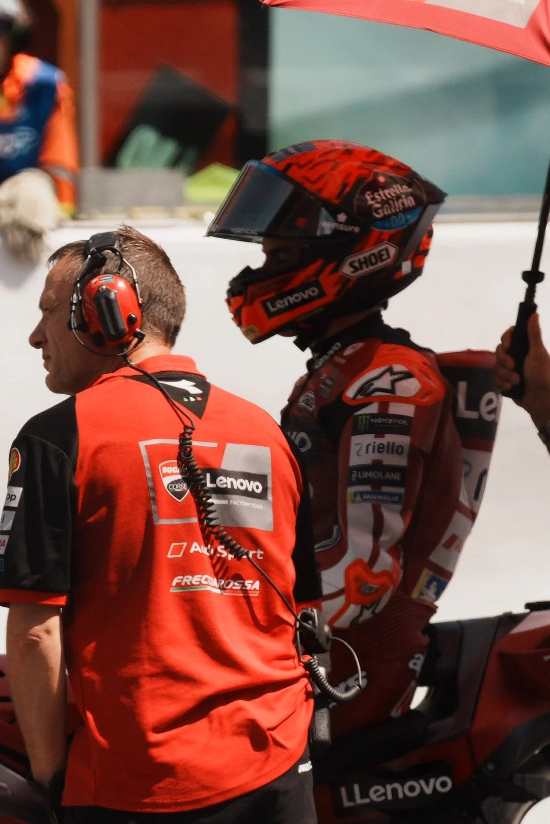 Motorsport team members in red and black uniforms preparing for a race, with one figure wearing a red helmet and holding an umbrella, amidst a racing setting.