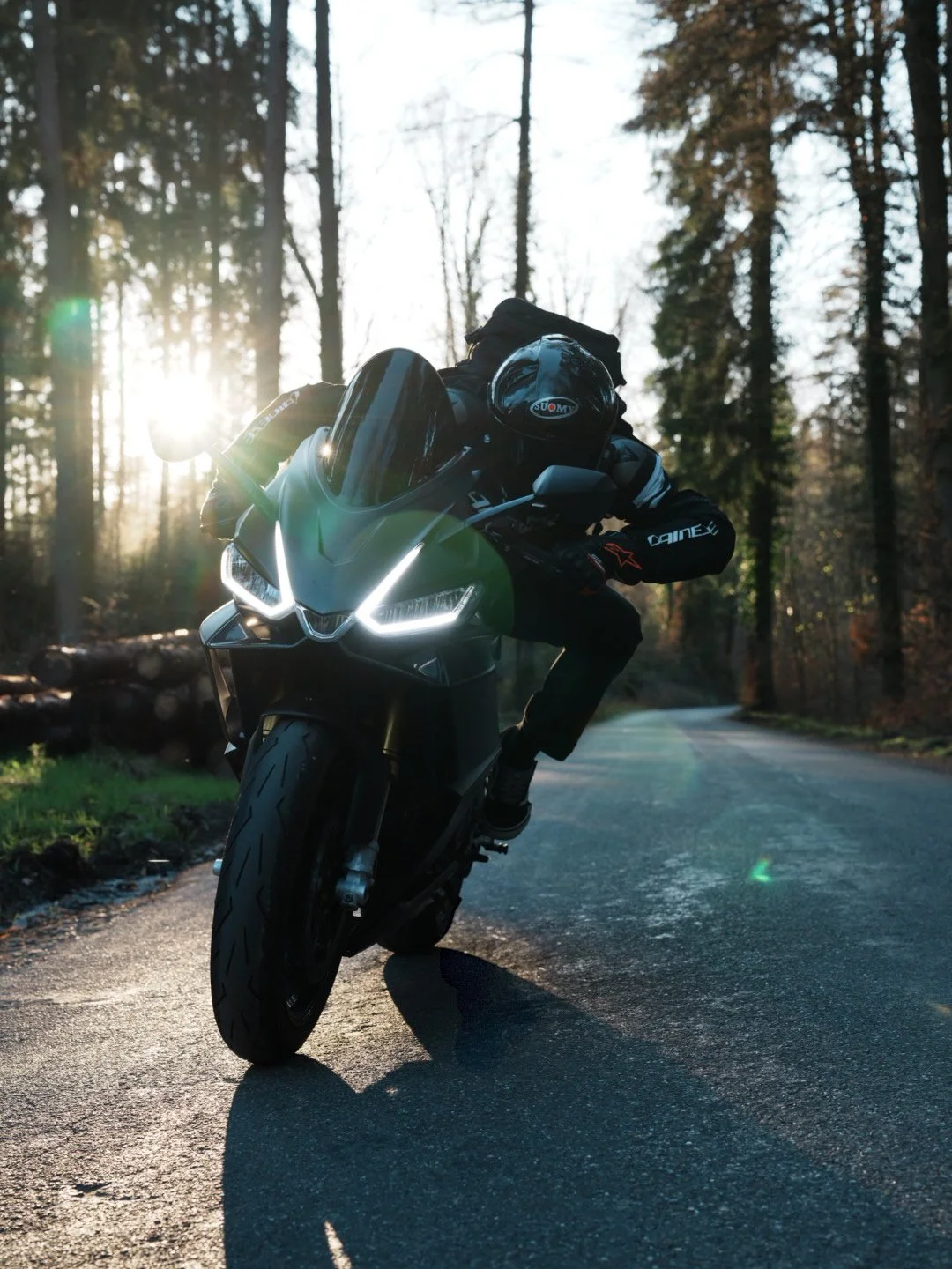 Motorcyclist riding on a forest road during sunset, leaning into a turn with the sun filtering through tall trees.