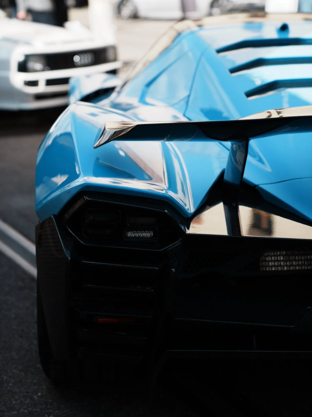 Close-up of the front of a blue luxury sports car with sharp angular design, parked outdoors with other vehicles in the background.