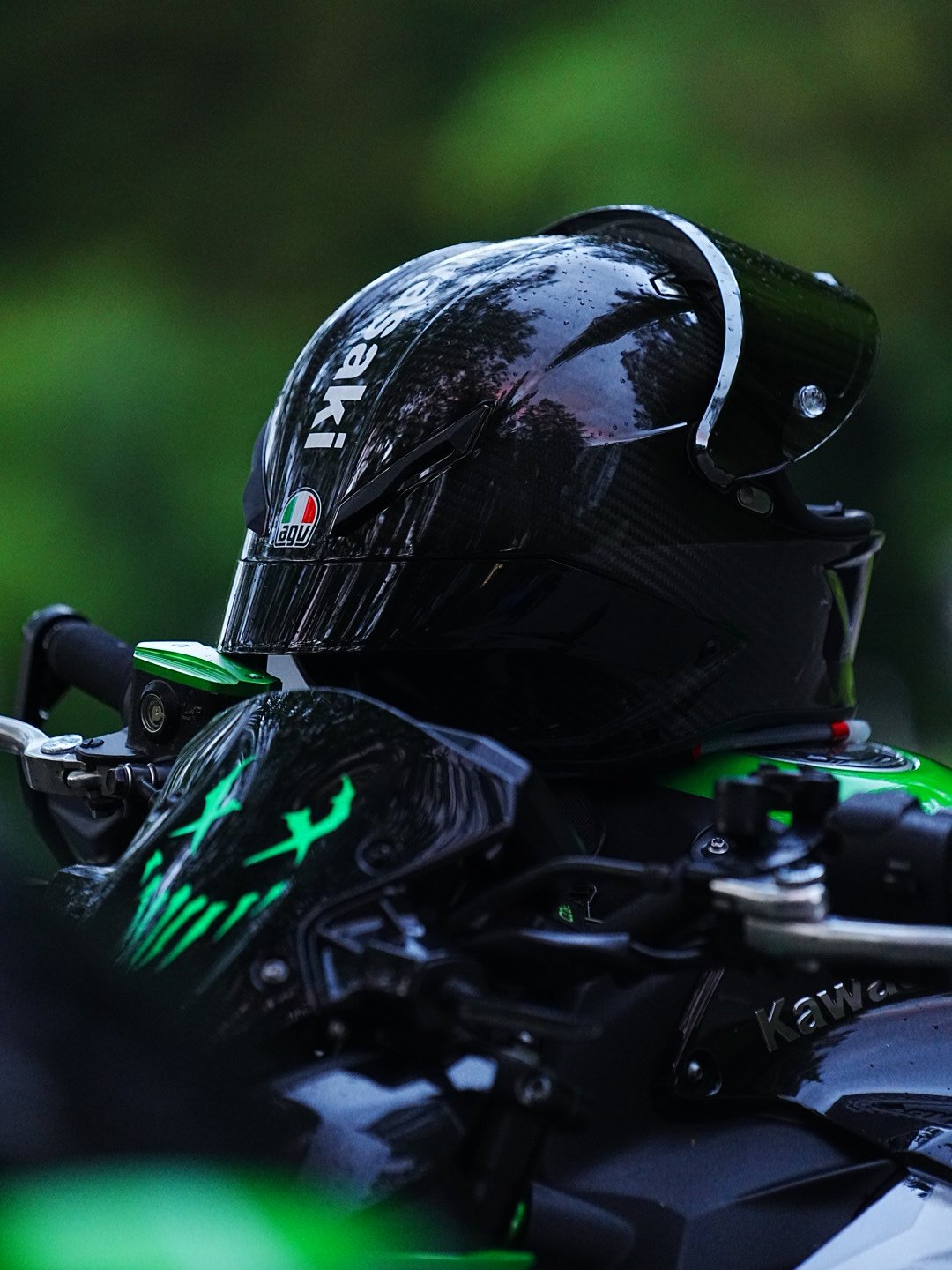 A black motorcycle helmet with a glossy finish placed on a sports motorcycle. The helmet bears the AGV logo, and the motorcycle features a Monster Kawasaki logo with green accents and is outdoors with a blurred green background.