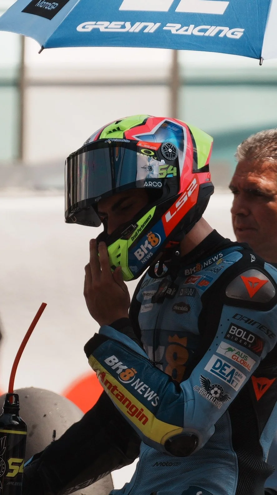 A motorcycle racer in racing gear, wearing a colorful helmet, sitting under a blue umbrella with the GRESINI RACING logo.