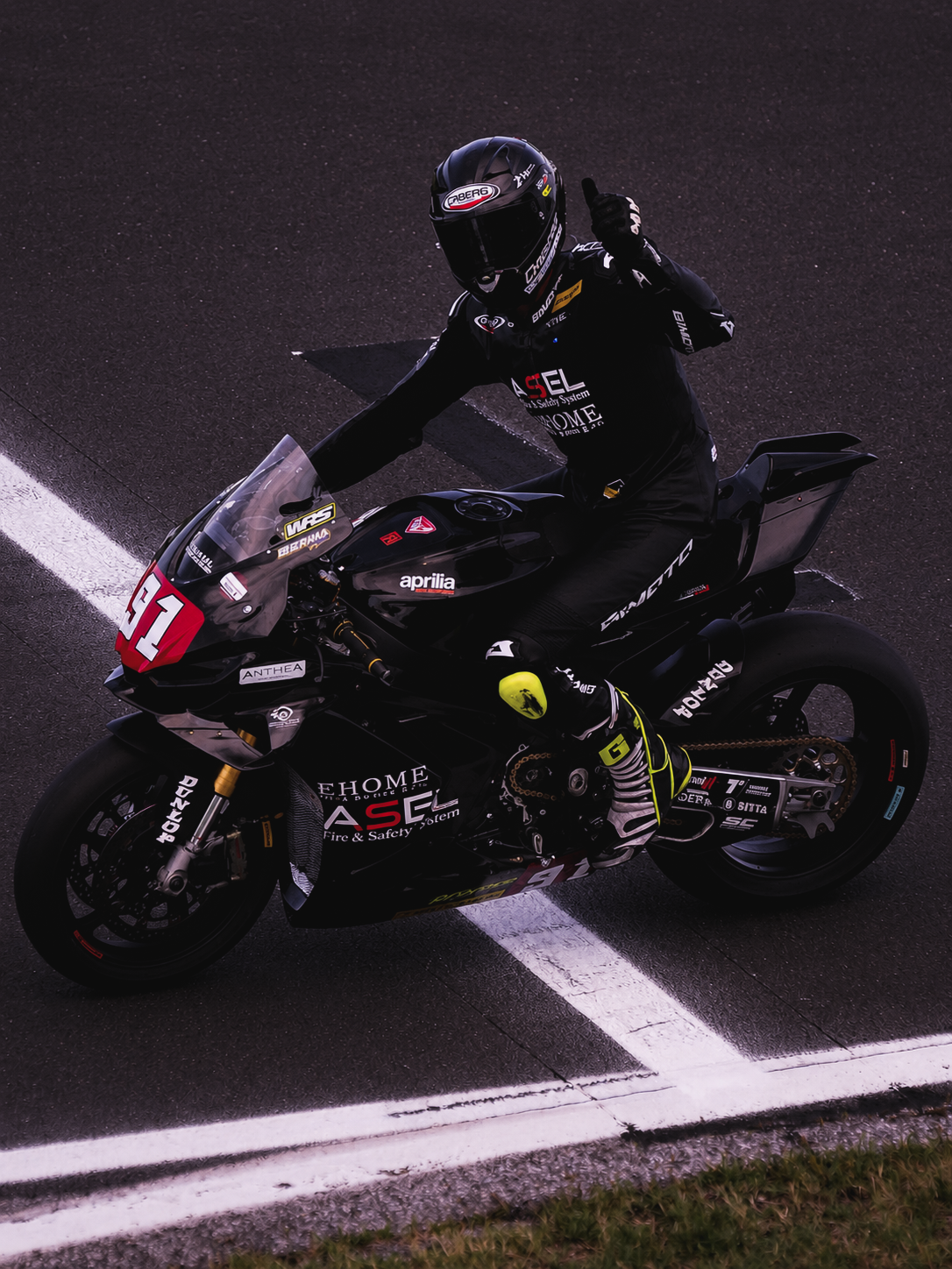Motorcycle racer on a black Aprilia bike, wearing black racing gear and helmet, giving a thumbs-up, on a race track.
