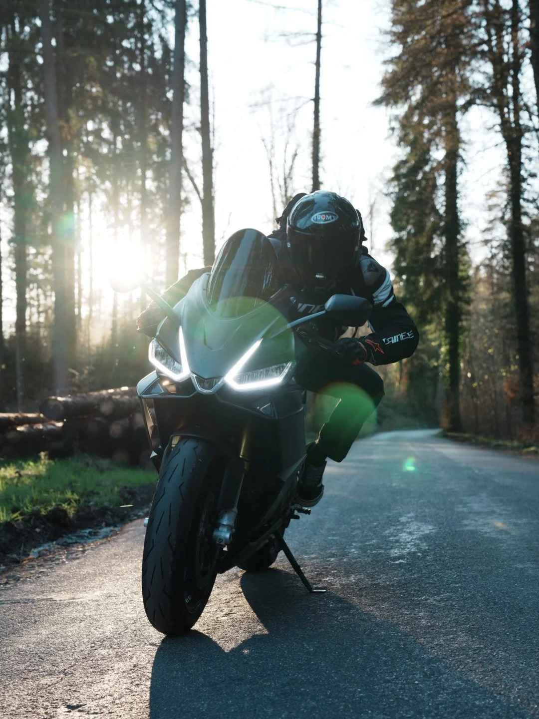 Motorcyclist in black gear riding a black motorcycle on a forested road with sunlight filtering through trees.
