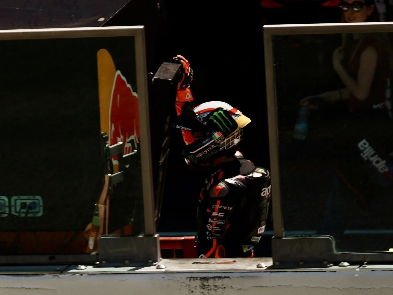Motorcycle racer wearing a helmet and racing suit standing in a garage area, with two glass panels in front showing the scene.