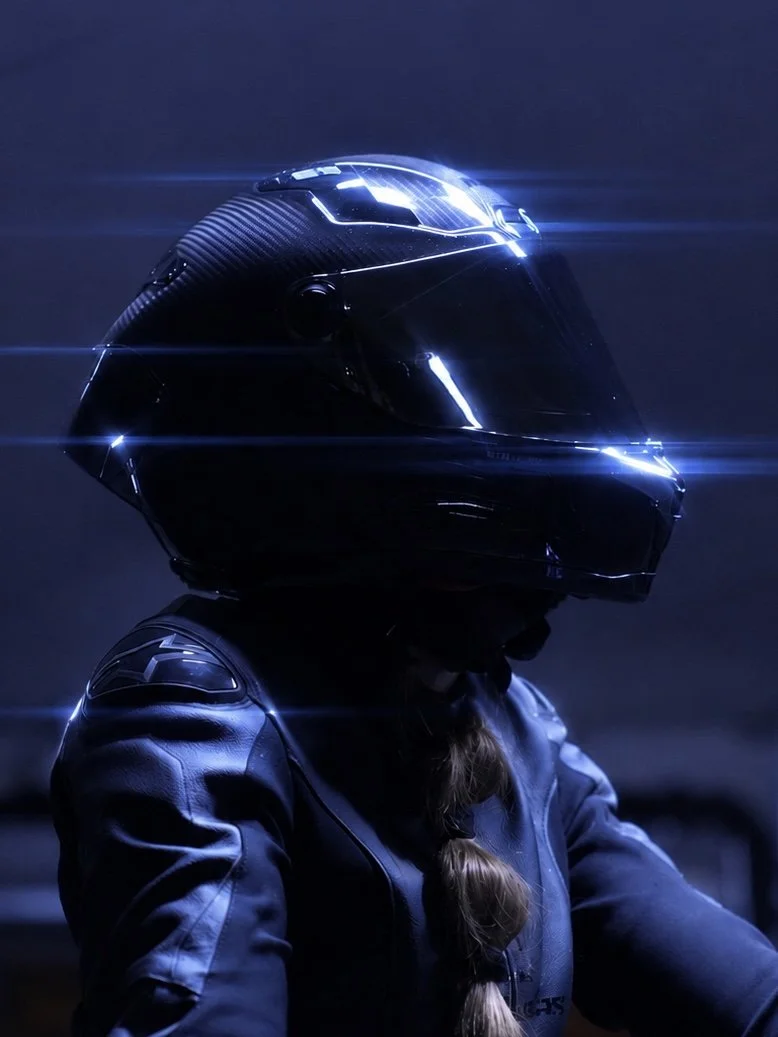 Person wearing a black racing helmet and a blue racing suit, with long hair in a braid, in a dark setting with reflective light lines.