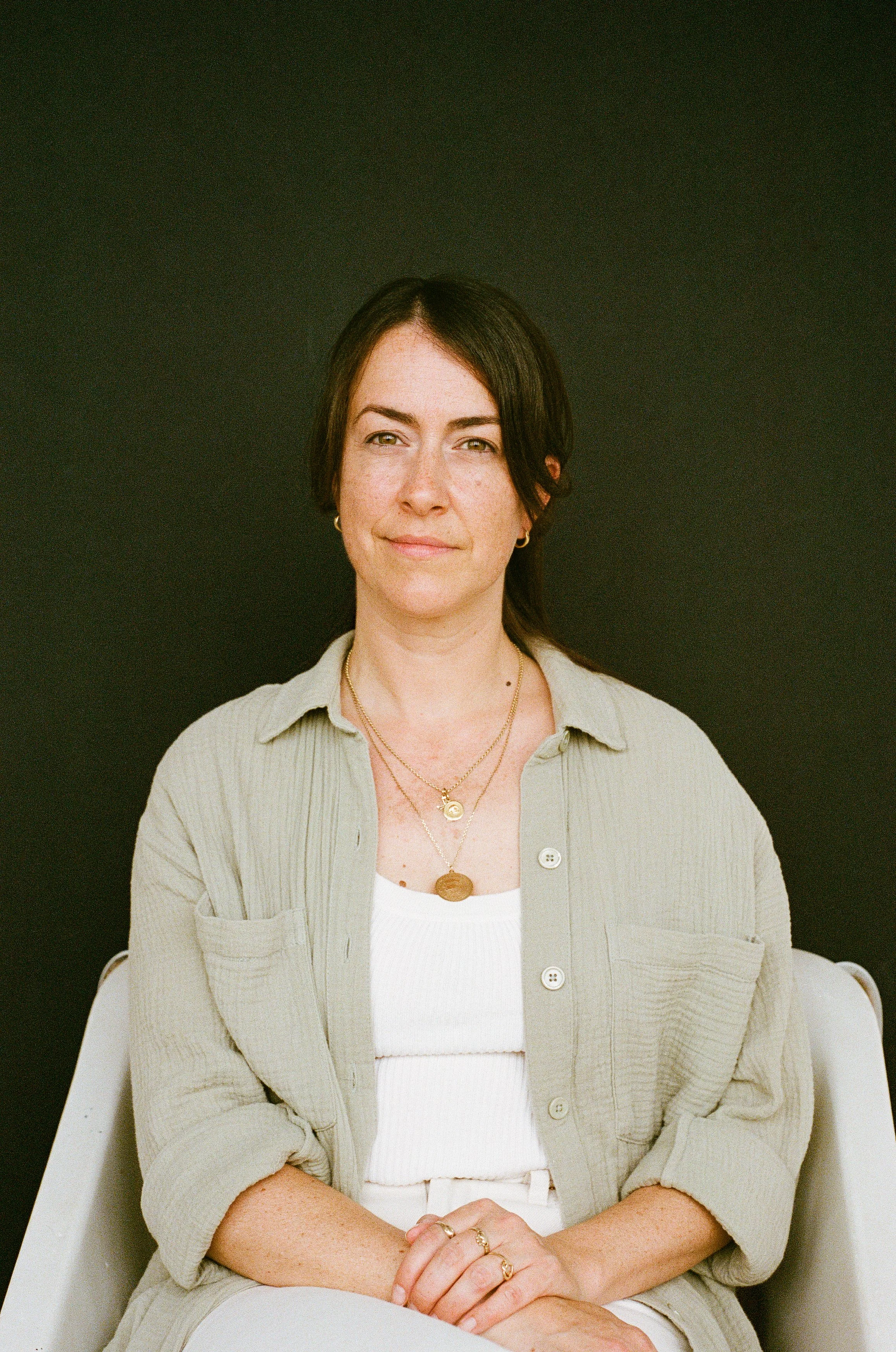 Une femme avec des cheveux bruns courts, portant une chemise beige avec des boutons, des colliers en or et des bagues, assise sur une chaise blanche contre un fond noir.