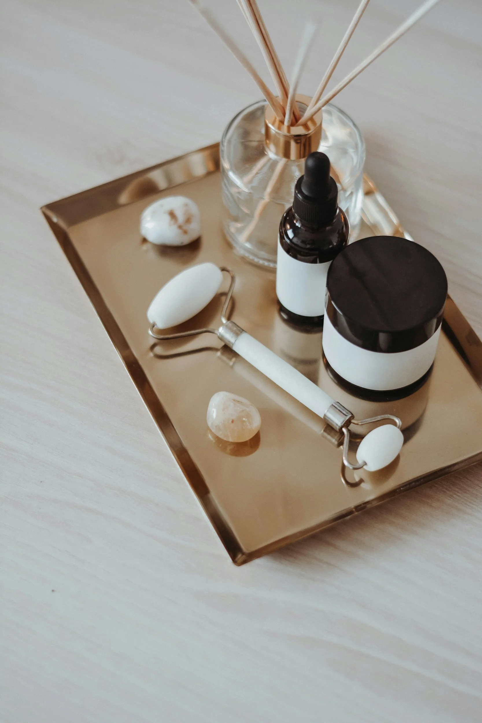 A gold tray with a glass bottle filled with reed diffuser sticks, a dark amber dropper bottle, two small jars, a white facial roller, and a white stone on a light silver surface.