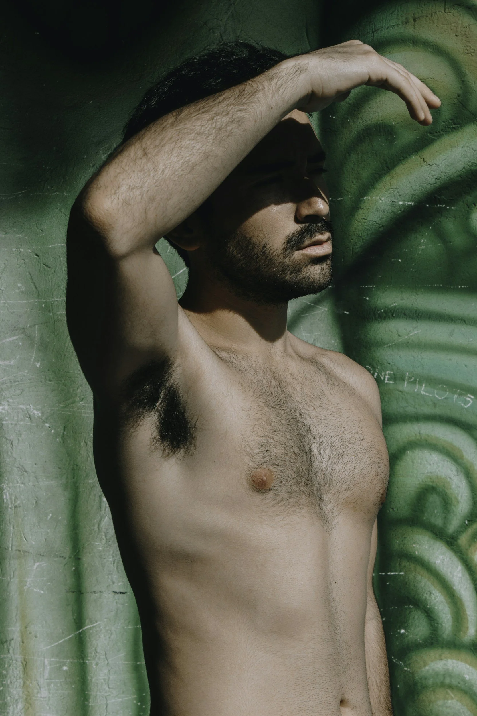 A shirtless man with a beard and dark hair, standing against a textured green wall, with one arm raised and hand shielding his eyes from sunlight.