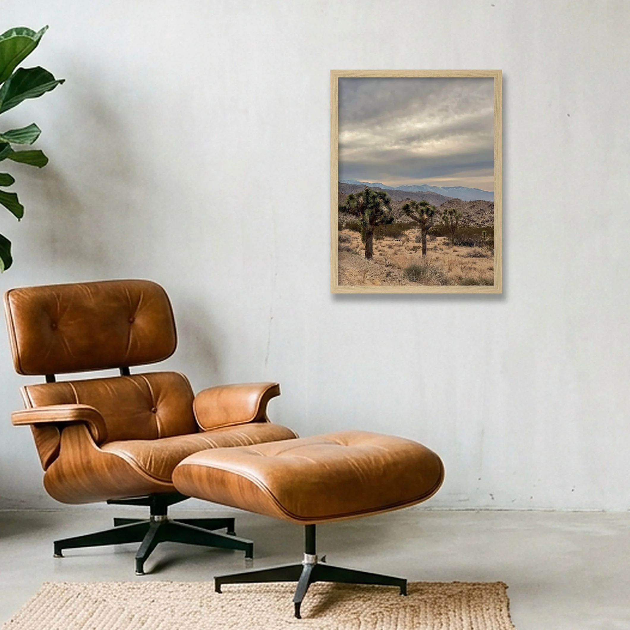 A light oak framed landscape print of a stormy desert vista hanging on a concrete wall above a modern leather lounge chair and green indoor plant.
