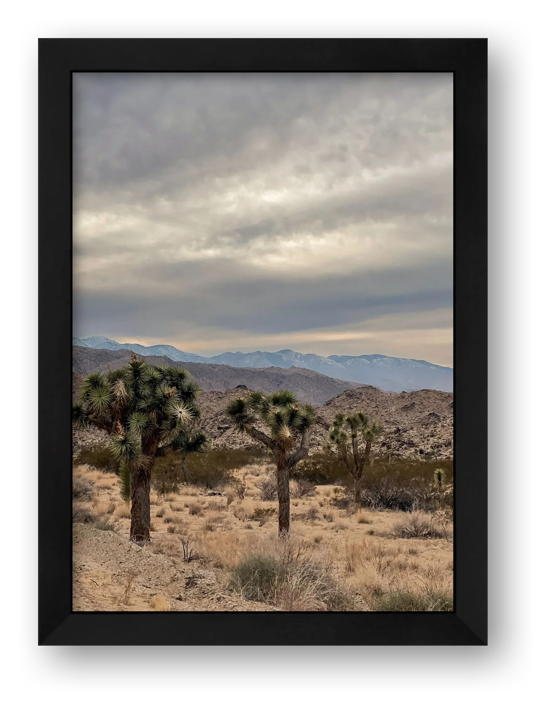 A black wooden gallery frame displaying a fine art print of dark storm clouds hovering over a high desert mountain landscape with a wide cream mat border.