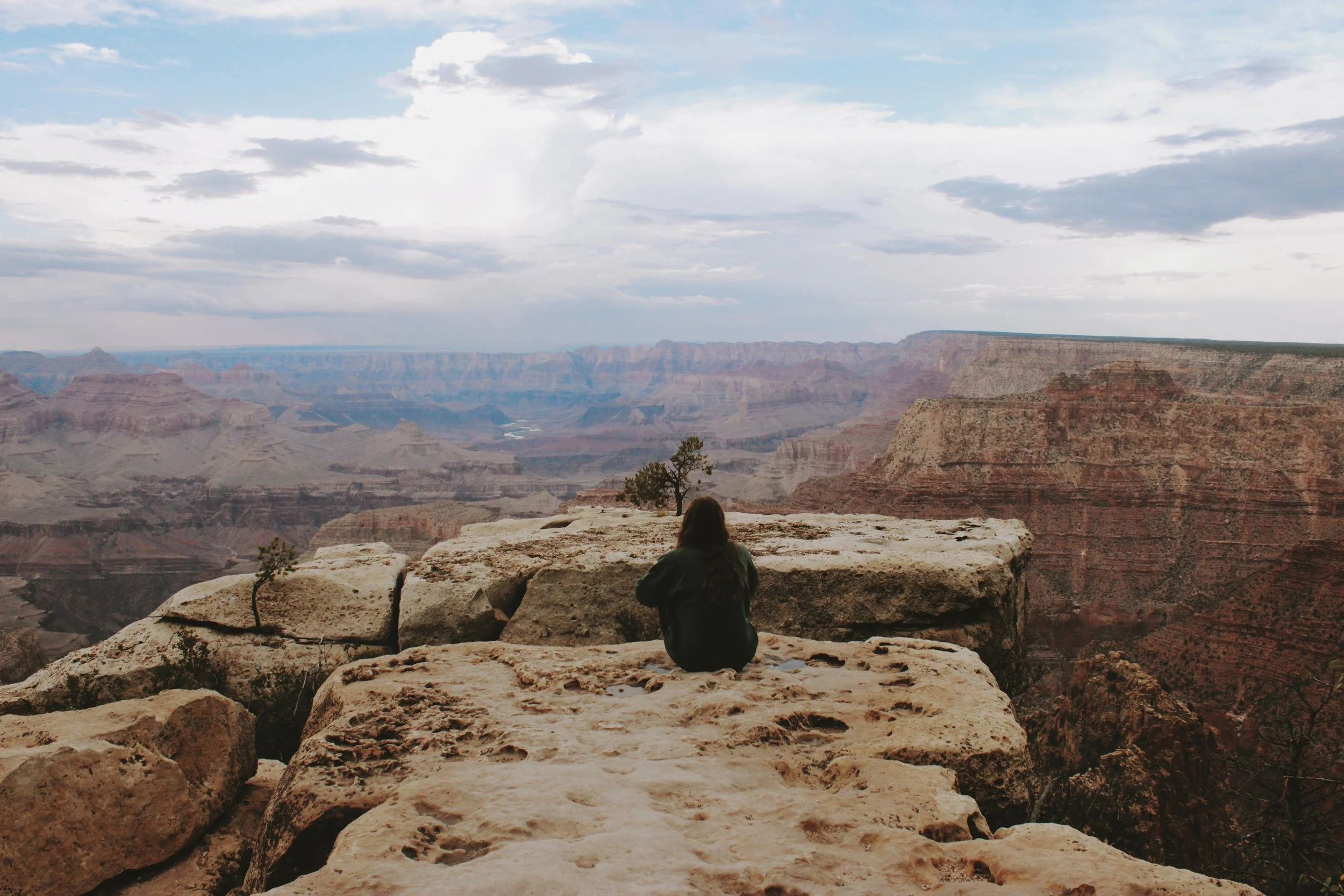 A person sitting on rocks at the Grand Canyon, looking at the expansive canyon and cloudy sky.
