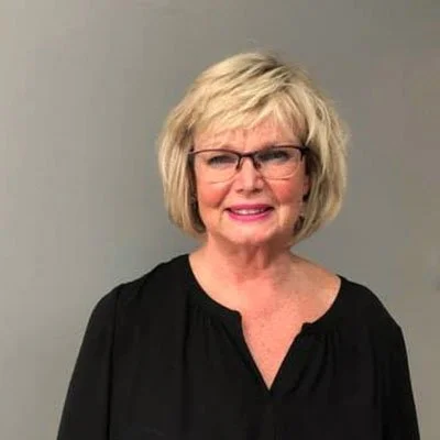 A smiling middle-aged woman with blonde hair, wearing glasses and a black blouse, standing against a plain gray background.