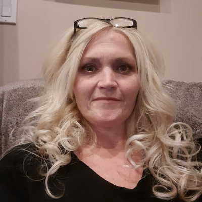 A woman with curly blonde hair, wearing glasses on top of her head, sits on a gray couch in a room with beige walls.