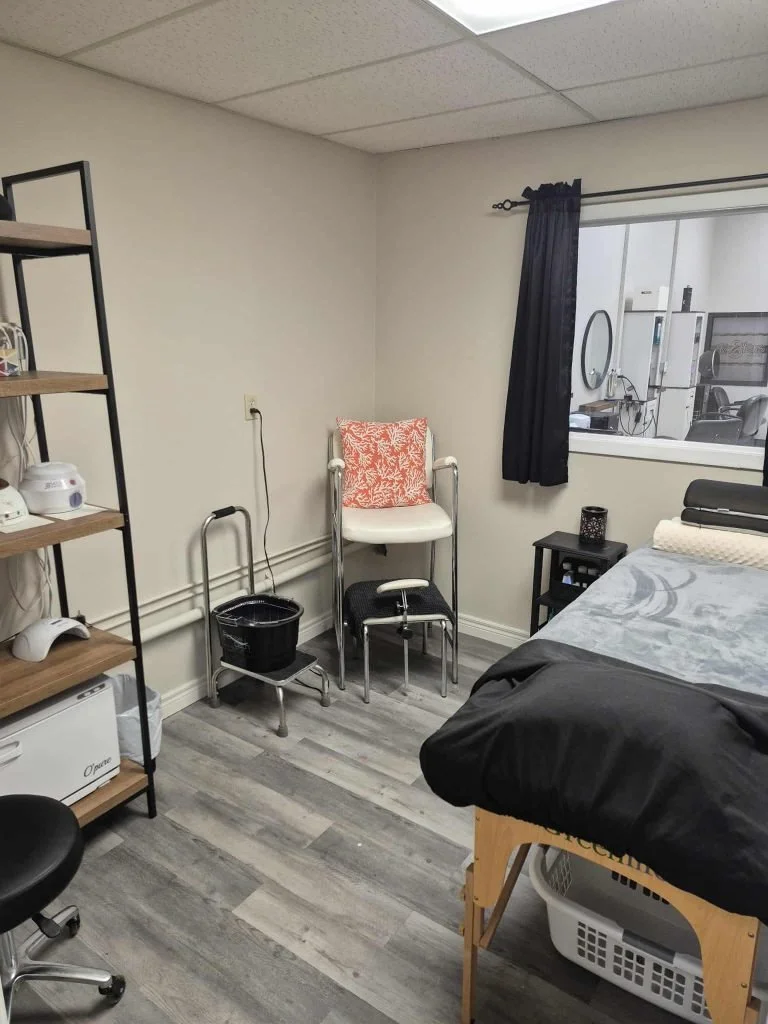 Medical or therapy room with a bed, black blanket, white pillow, small black side table, window with black curtains, chairs, a shelf with medical equipment, and a cart with supplies.