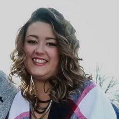 Smiling woman with shoulder-length wavy hair, wearing a multicolored sweater and layered necklaces.