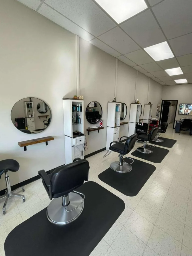 Empty hair salon with four styling stations, black chairs, mirrors, and supplies on shelves.