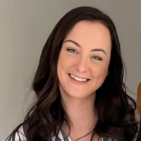 Smiling woman with long dark hair and fair skin, wearing a white top, against a neutral background.