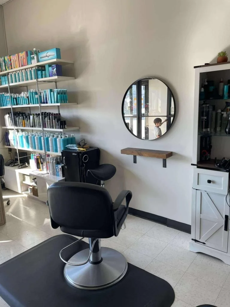 Inside a hair salon with a black styling chair on a black mat, shelving with hair products, a potted plant on top of a cabinet, and a round mirror on the wall reflecting a young boy.