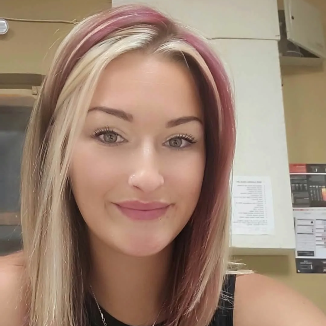 A young woman with shoulder-length blonde hair with pink highlights, smiling, in an indoor setting with a bulletin board and electrical panel in the background.