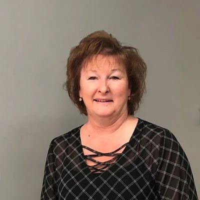 A woman with short brown hair wearing a black and gray checkered top standing against a gray wall.