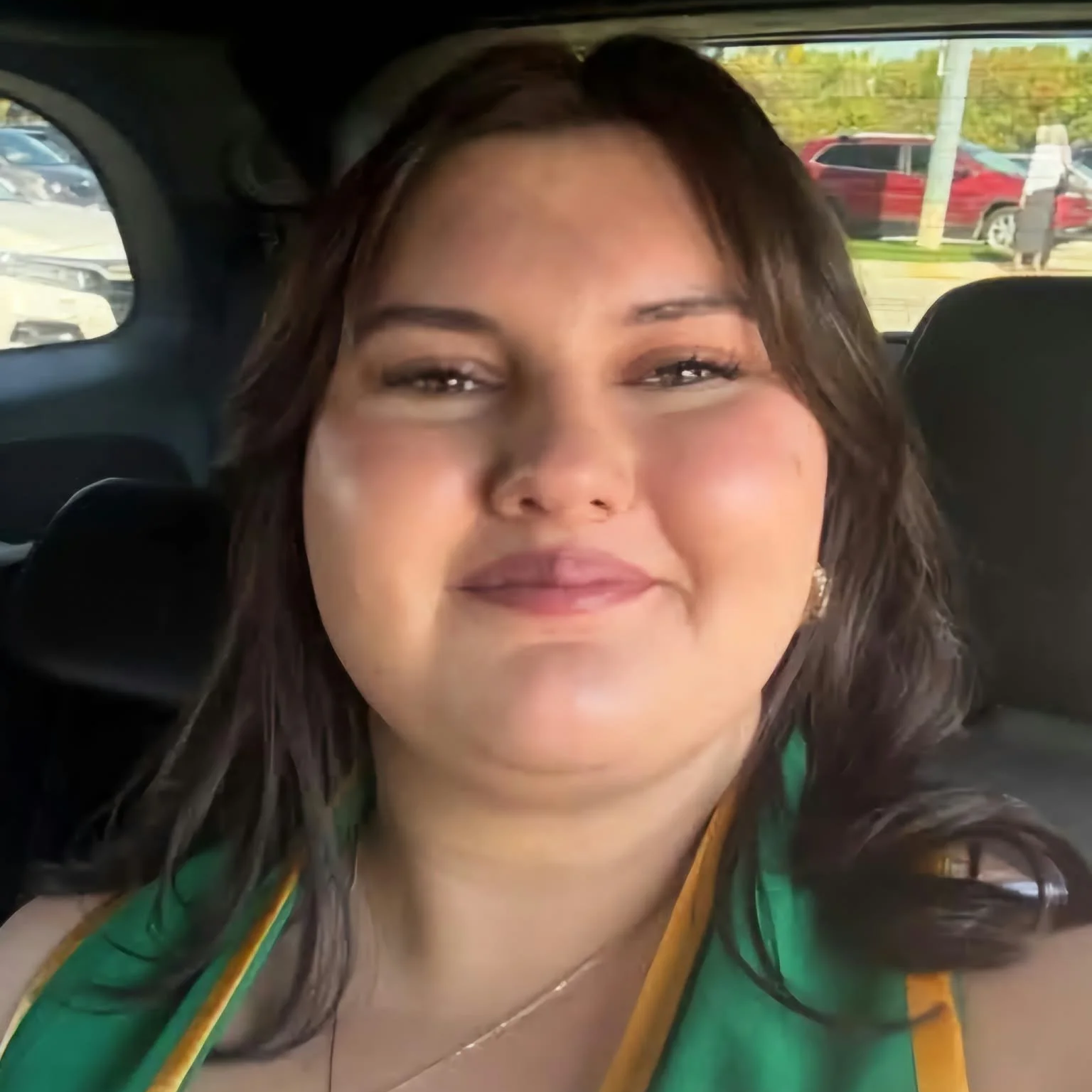 A woman with long dark hair, wearing a green and yellow outfit, smiling at the camera inside a vehicle with other cars and a person walking outside visible through the window in the background.