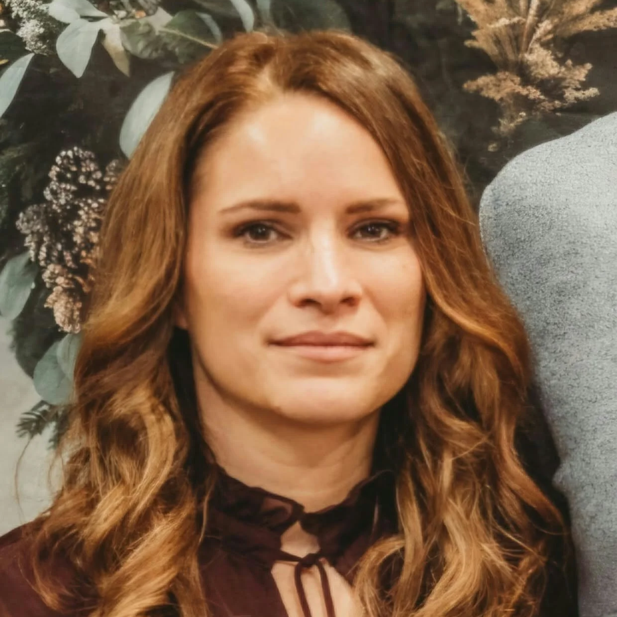 Close-up portrait of a woman with long auburn hair wearing a burgundy blouse, posed against a floral backdrop.