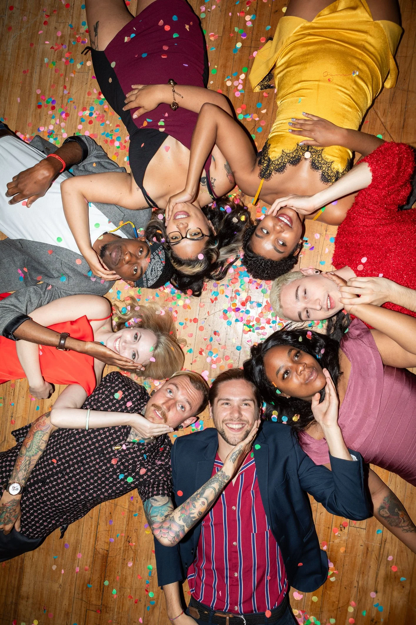 People lying together on a wooden floor amid colorful confetti.