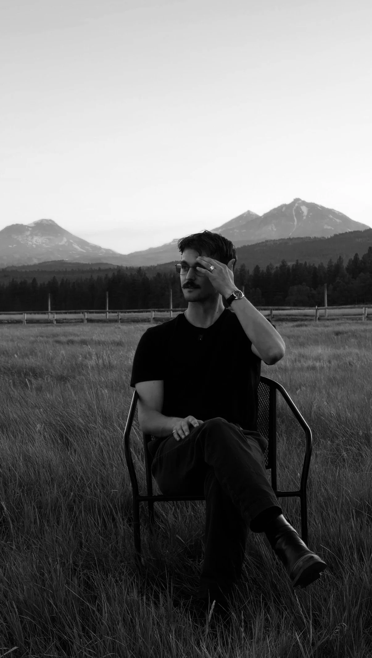 A man with glasses, a mustache, and dark hair sits on a chair in a field of tall grass, with mountains in the background. The photo is in black and white.