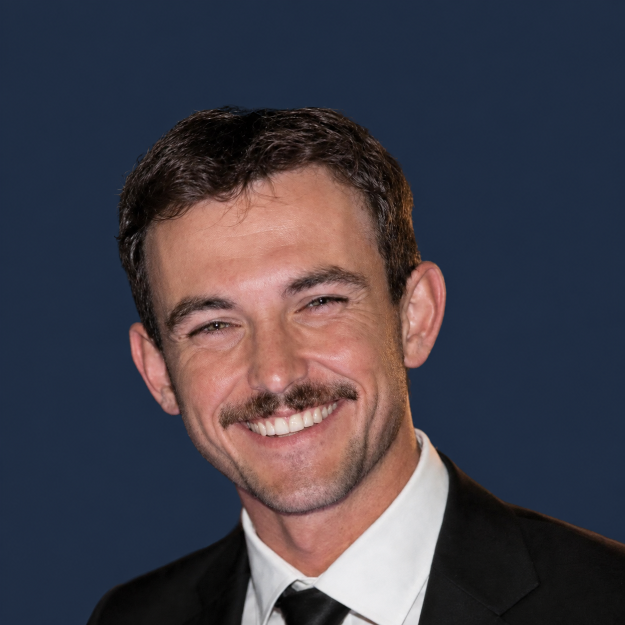 Smiling man in a suit and tie with a dark background.