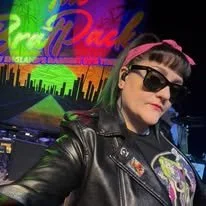 Person wearing black sunglasses and a black leather jacket with patches, standing in front of a colorful neon sign that says 'Pink' in an entertainment venue.