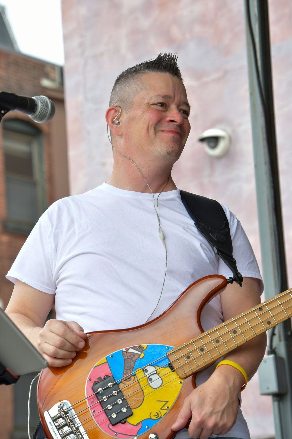 A man with a mohawk hairstyle and earrings playing a colorful bass guitar, smiling and wearing a white t-shirt, in an outdoor setting.