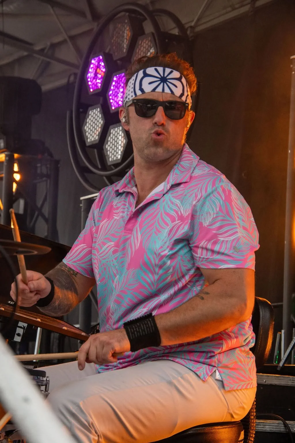 Man wearing a blue and white bandana, black sunglasses, pink and blue patterned shirt, and beige pants playing drums at a concert with colorful stage lighting.