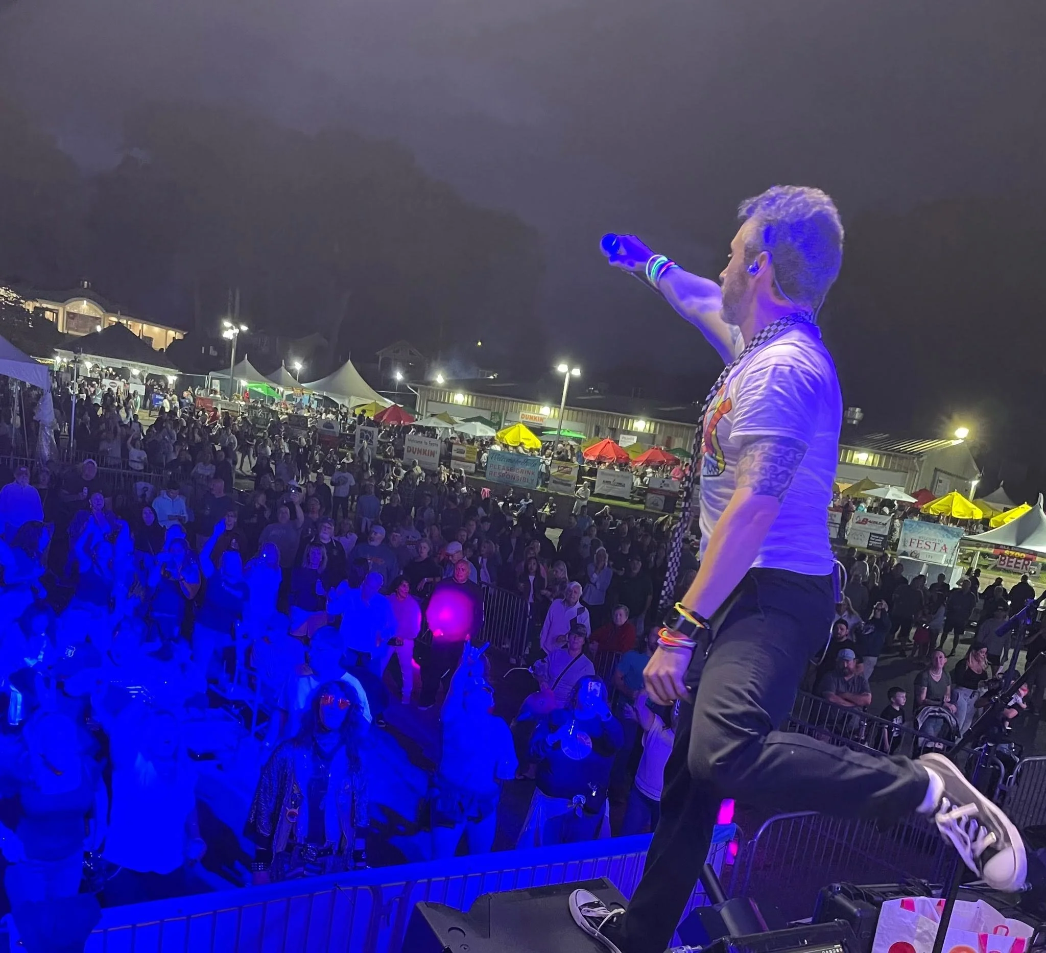 A performer on stage at night with their arm raised, illuminated by purple-blue lighting, in front of an audience at an outdoor event.