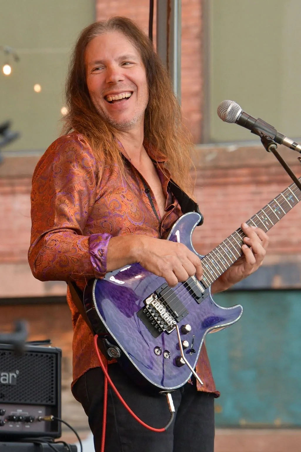 A man with long hair, wearing a patterned shirt, playing an electric guitar and smiling during a performance.