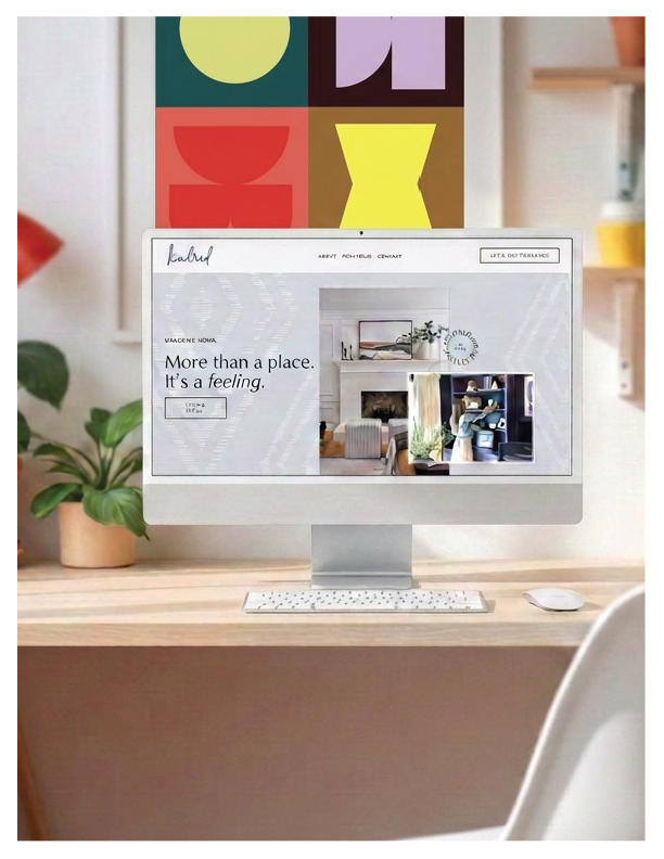 An Apple iMac computer on a wooden desk with a small potted plant on the left and colorful abstract art hanging on the wall behind it.