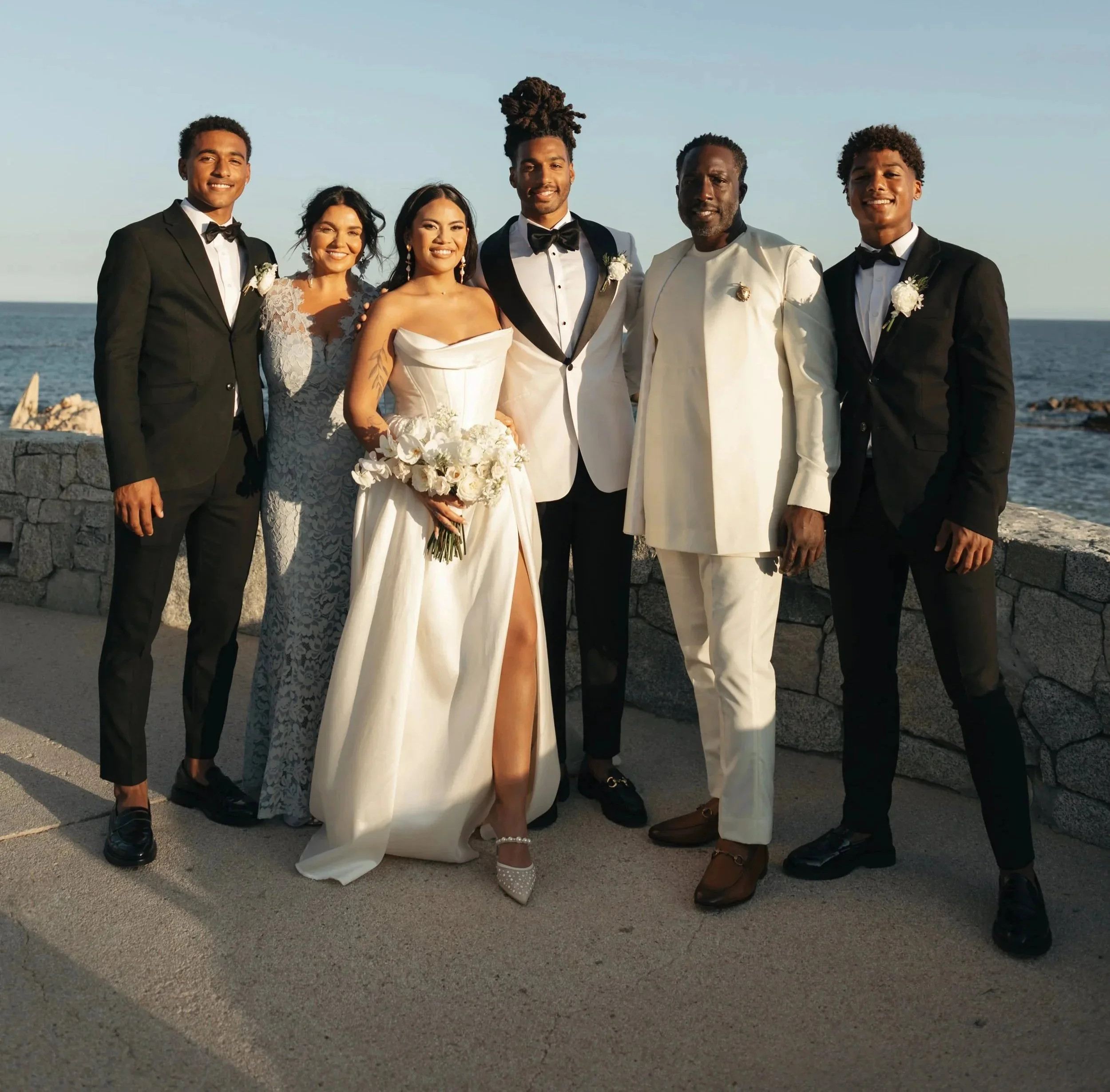 Group of six people dressed formally, at a wedding, standing outdoors by the sea, smiling for the photo.
