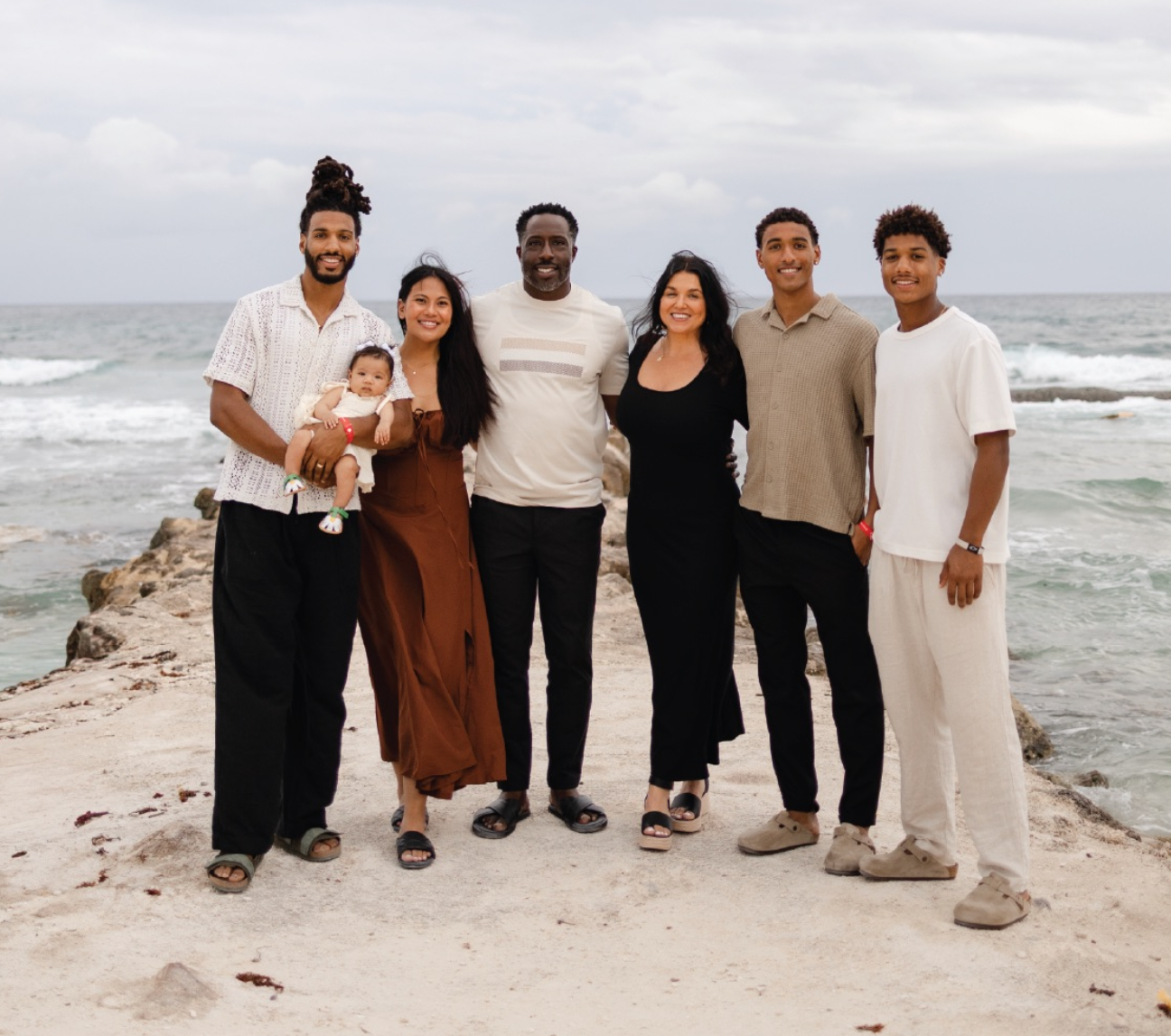 A diverse family of seven standing on a rocky beach with the ocean and cloudy sky in the background, smiling at the camera.
