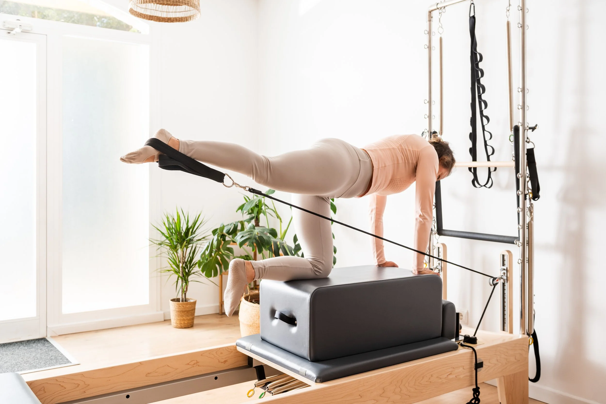 A woman is doing a Pilates exercise in a bright, modern studio using a reformer machine and resistance bands attached to a wall.