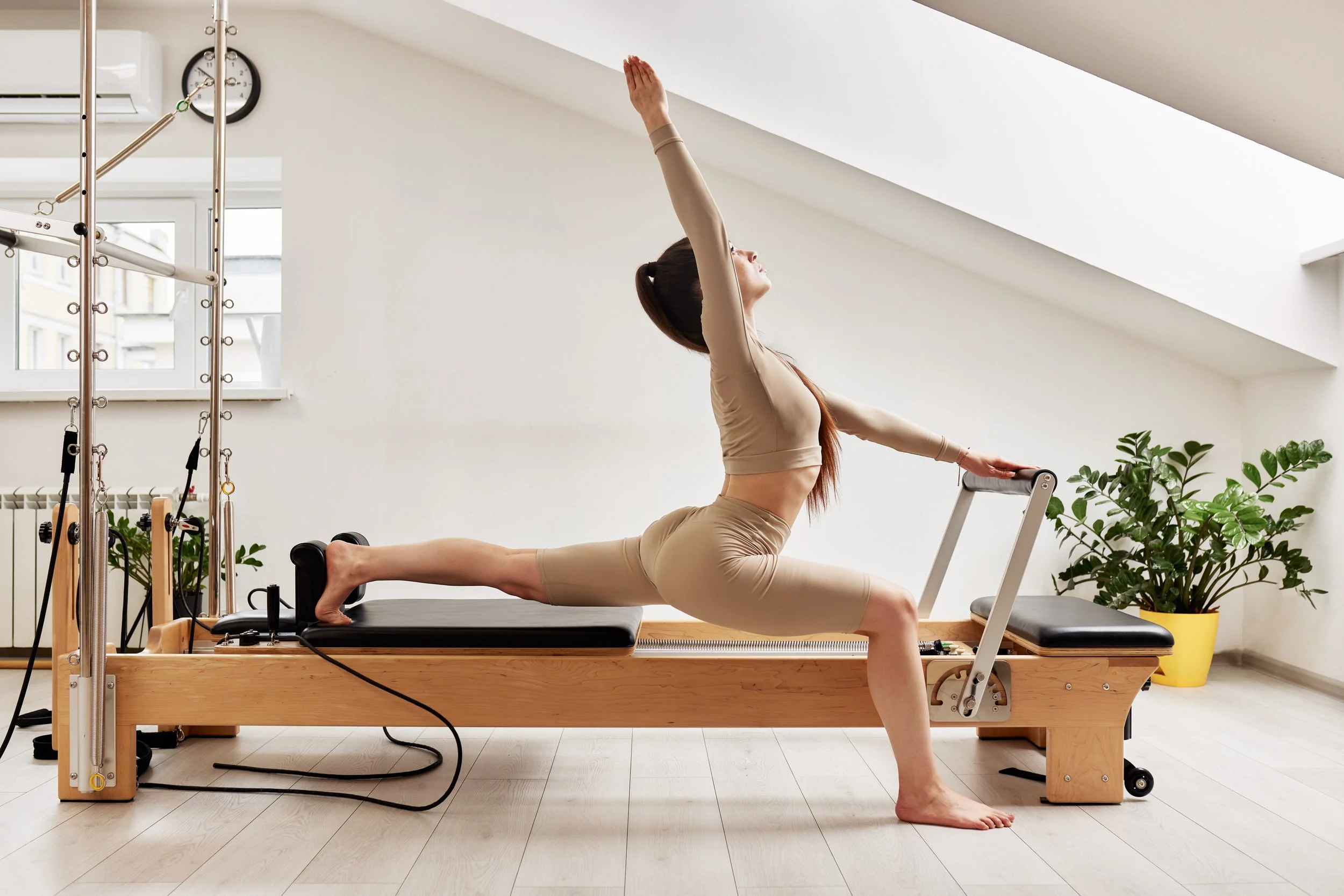 A woman practicing yoga on a Pilates reformer in a bright, minimalistic room with large windows, a wall clock, and a potted plant.