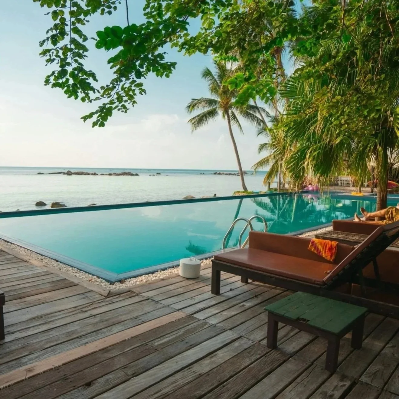 Swimming pool near the ocean with palm trees, lounge chairs, and a view of the sea and rocks in the background.