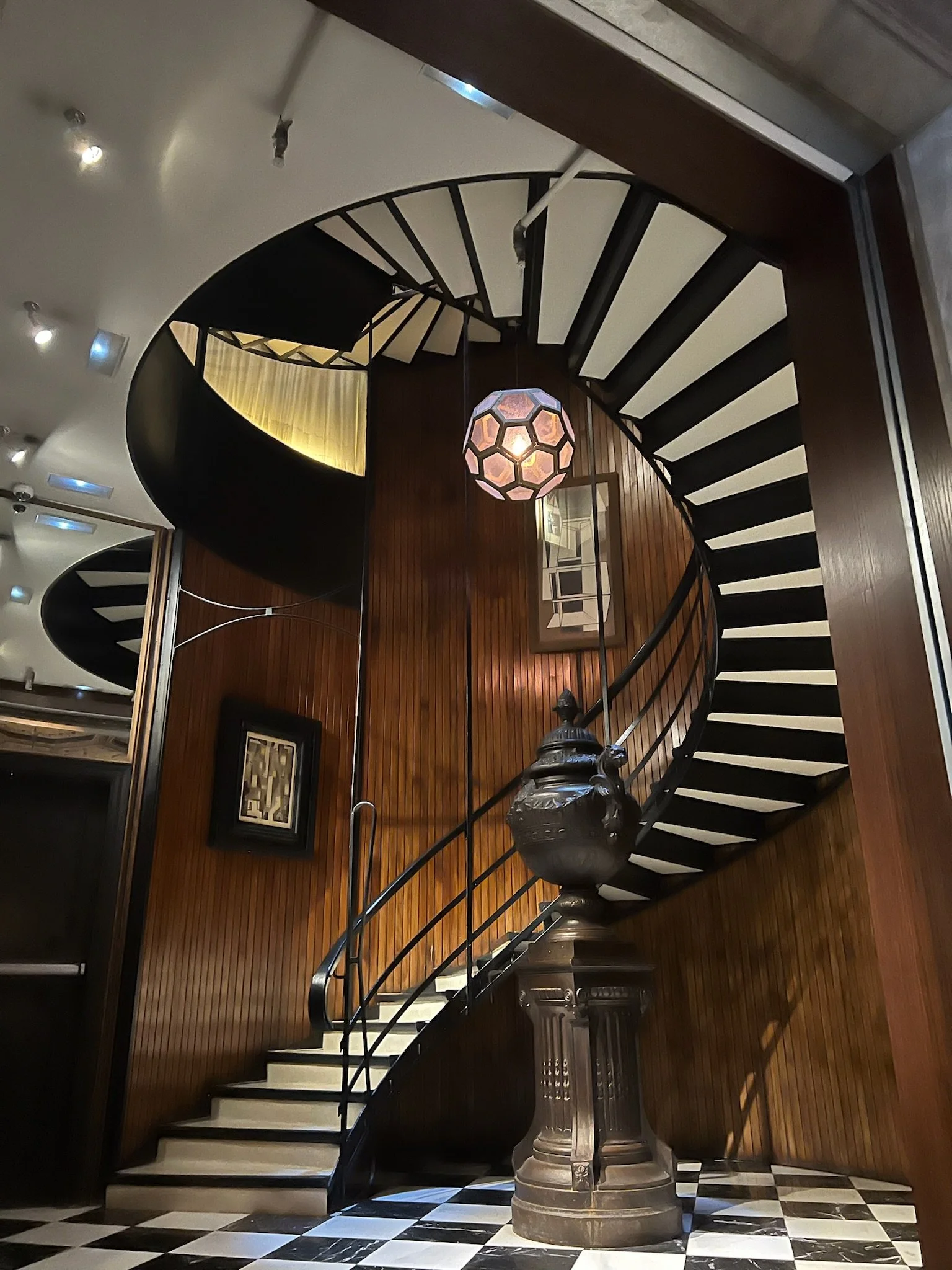 Interior of a building with a spiral staircase, a hanging hexagonal-shaped light fixture, wooden wall paneling, framed artwork, and a checkered black and white marble floor.