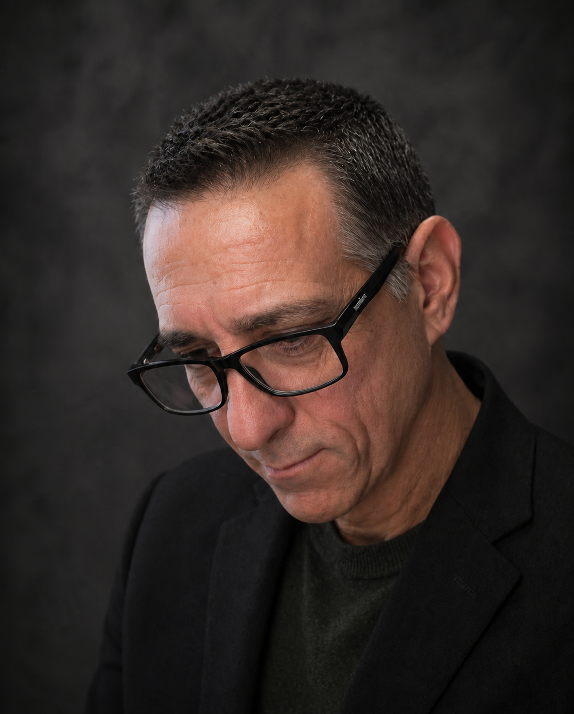 Kirk Saavedra a middle-aged man with short, dark, and slightly graying hair wearing black-rimmed glasses, a black blazer, and a dark shirt, looking down with a serious expression against a dark background.