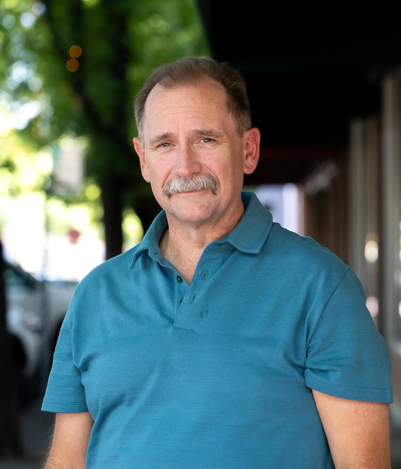 A middle-aged man with a mustache and short brown hair, wearing a blue polo shirt, standing outdoors with trees and a building in the background.