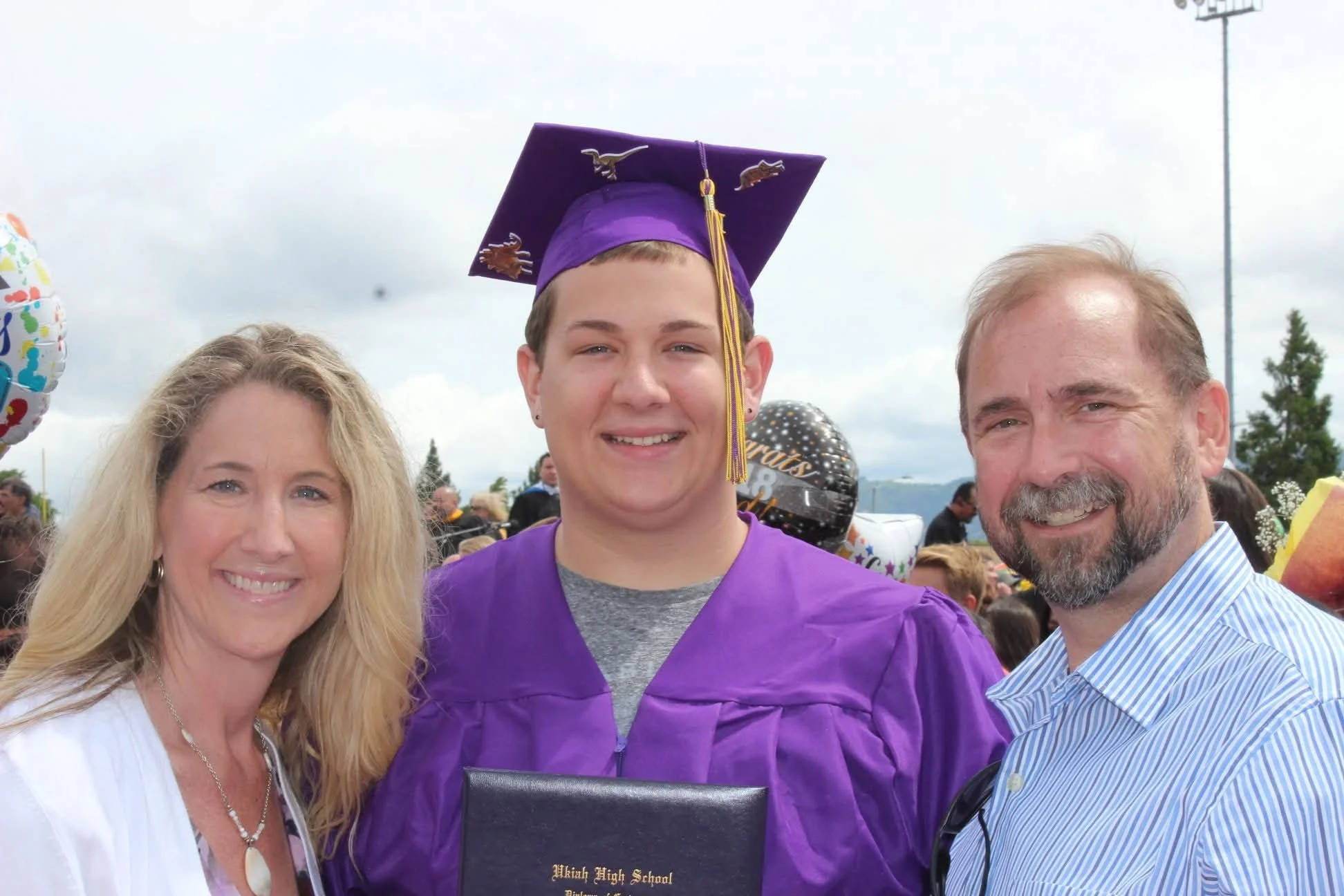 A young man wearing a purple graduation cap and gown, standing between a woman with long blonde hair and a man with a beard, both smiling at a graduation celebration outdoors.