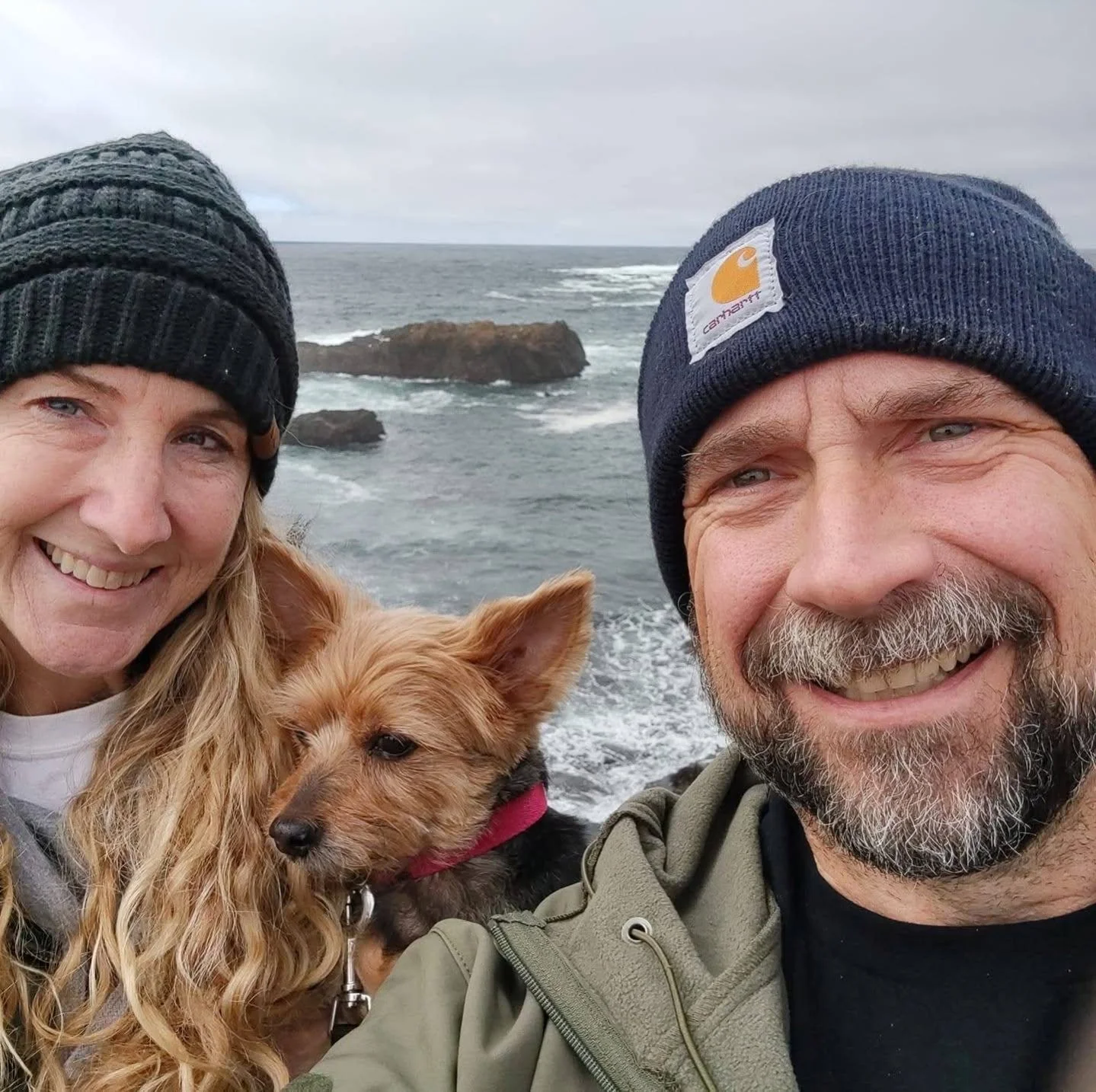 A smiling woman and man with a dog at the beach, ocean rocks in the background, both wearing winter hats.