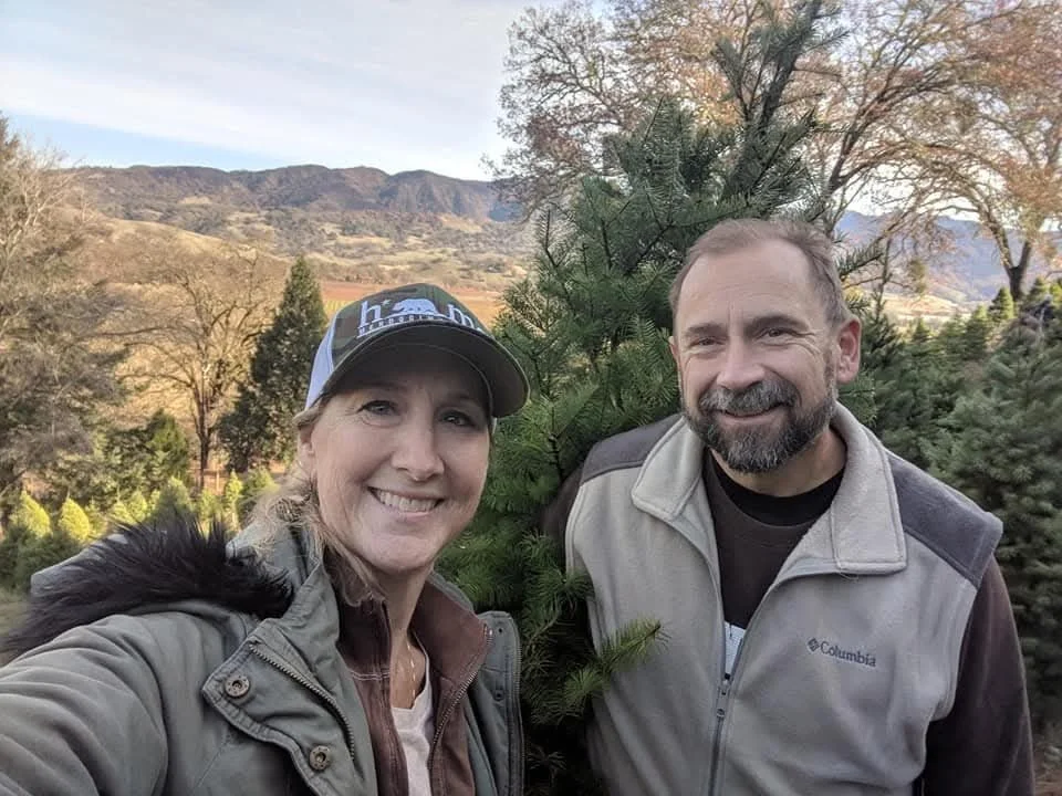 A smiling woman and man posing outdoors near a Christmas tree, with mountains and trees in the background.