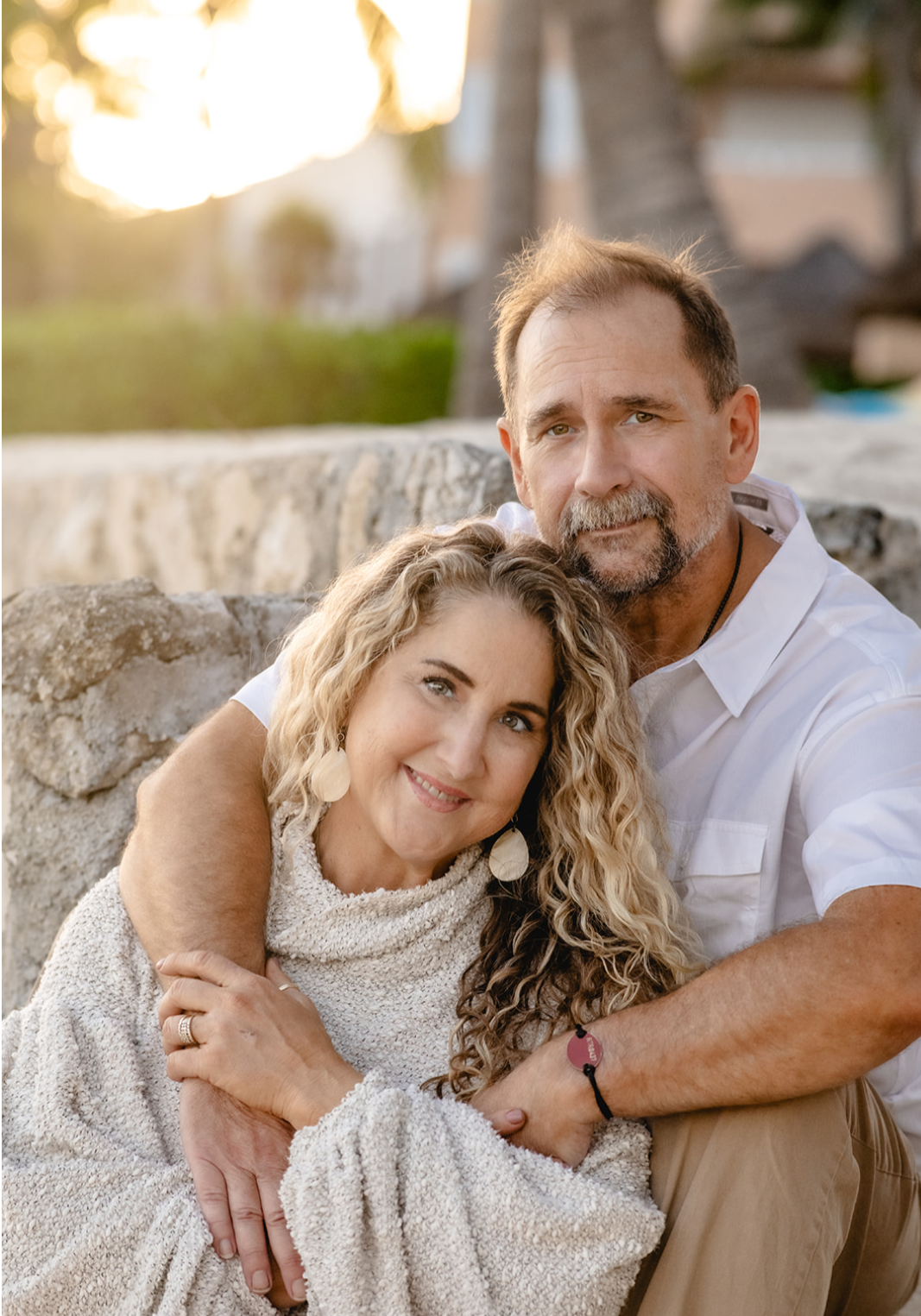 A couple sitting outdoors near a stone wall during sunset, hugging and smiling at the camera. The woman has long, curly blonde hair and the man has short brown hair and a beard.