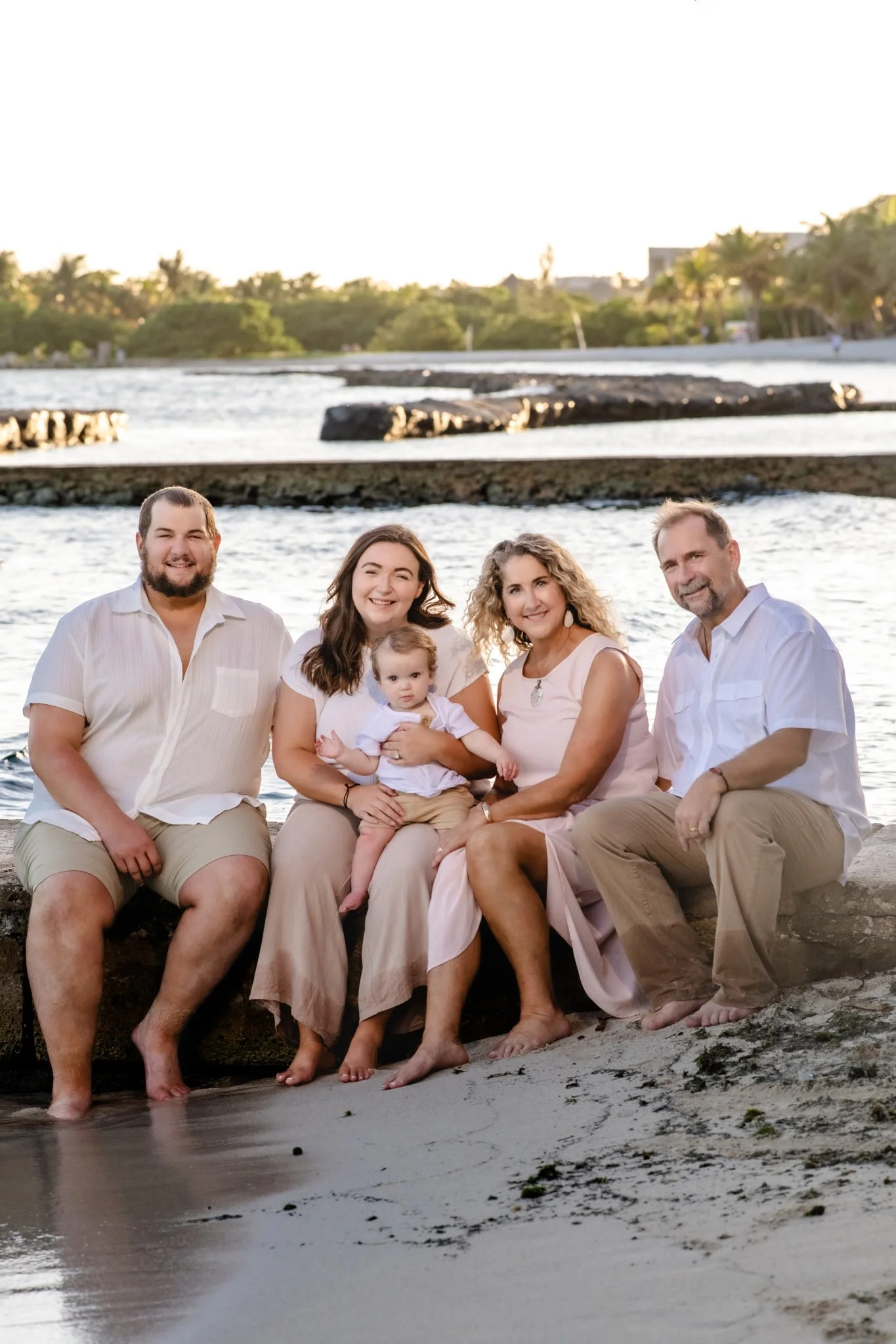 A family of five sitting on a beach at sunset. They are smiling, and there is water, sand, and trees in the background.