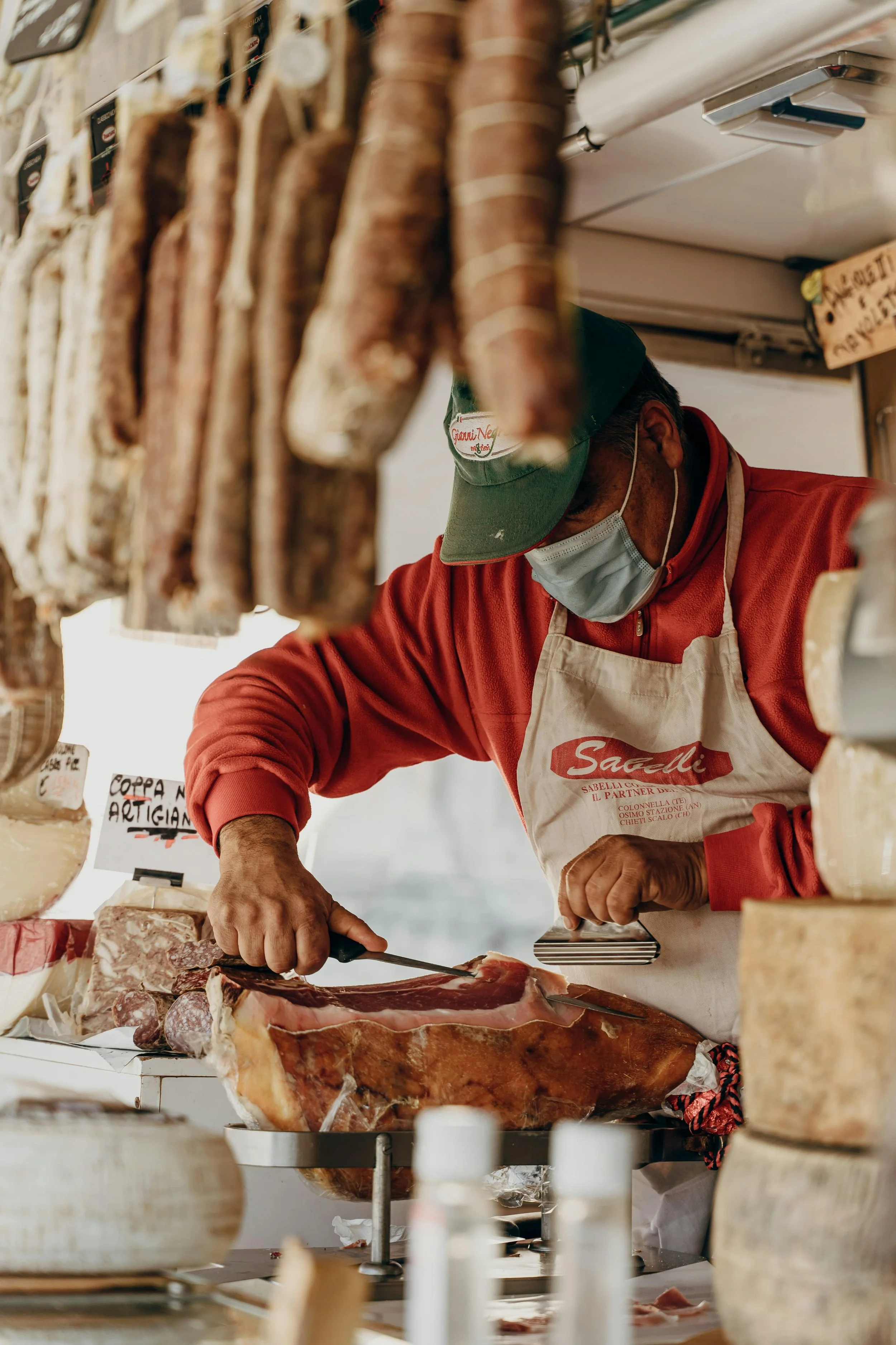 A man in a red jacket, green cap, and face mask is slicing cured ham at a cheese and deli shop, with hanging sausages and wheels of cheese visible around him.