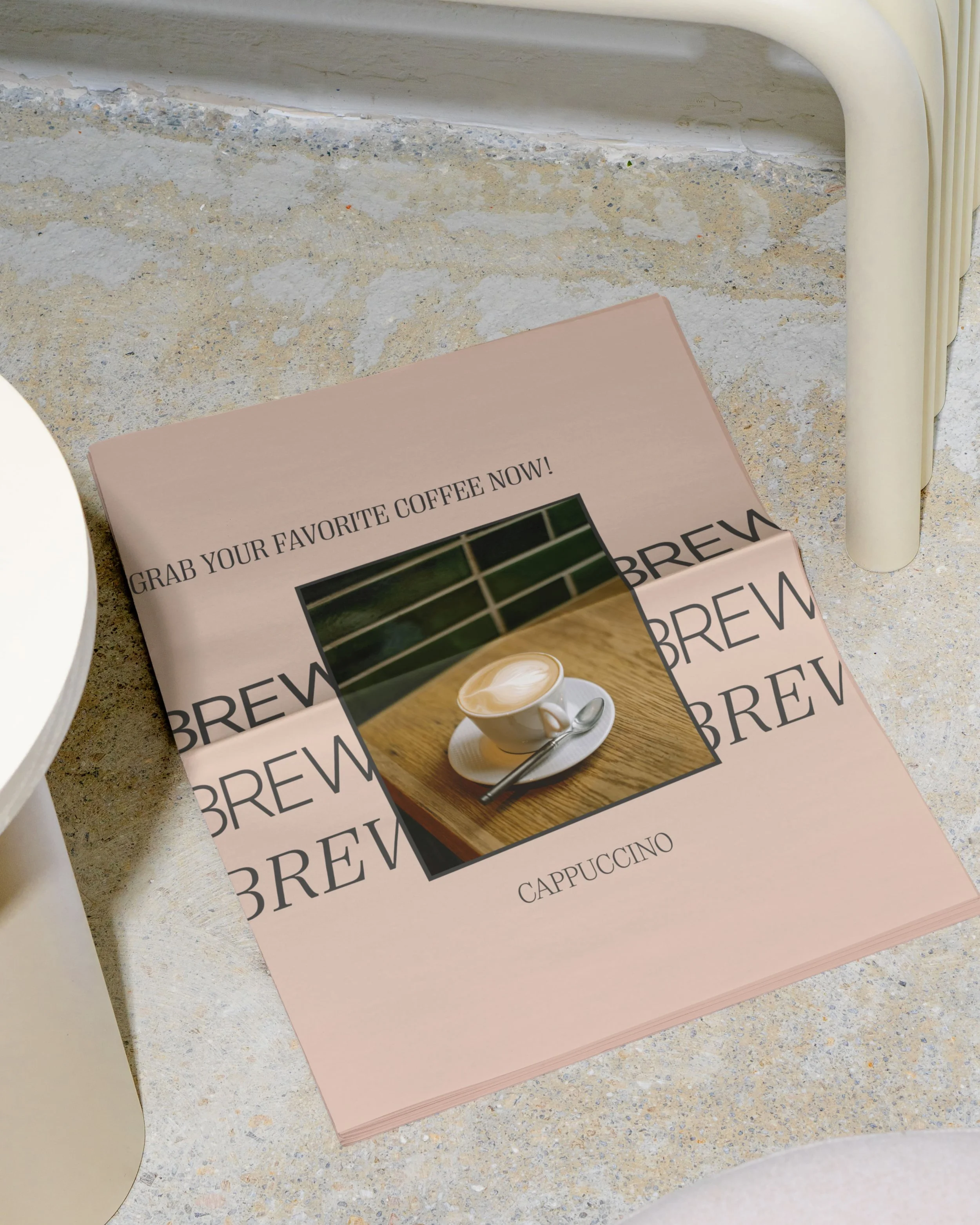 A magazine or flyer spread out on a speckled marble or concrete surface, featuring an image of a cup of cappuccino on a wooden table and the word 'BREW' repeatedly printed in the background with text inviting to grab coffee.